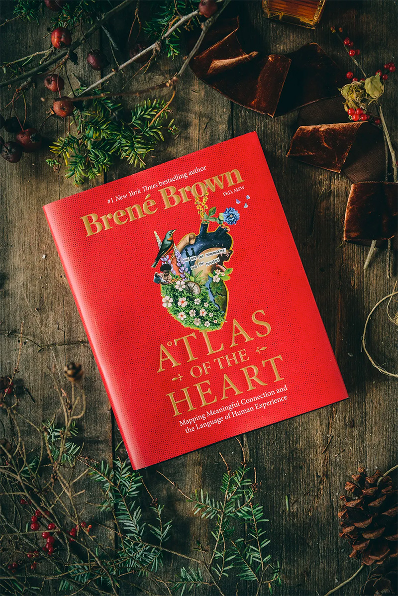 Rustic flatlay with pine, berries, and velvet ribbon. Red book cover that reads "Atlas of the Heart".