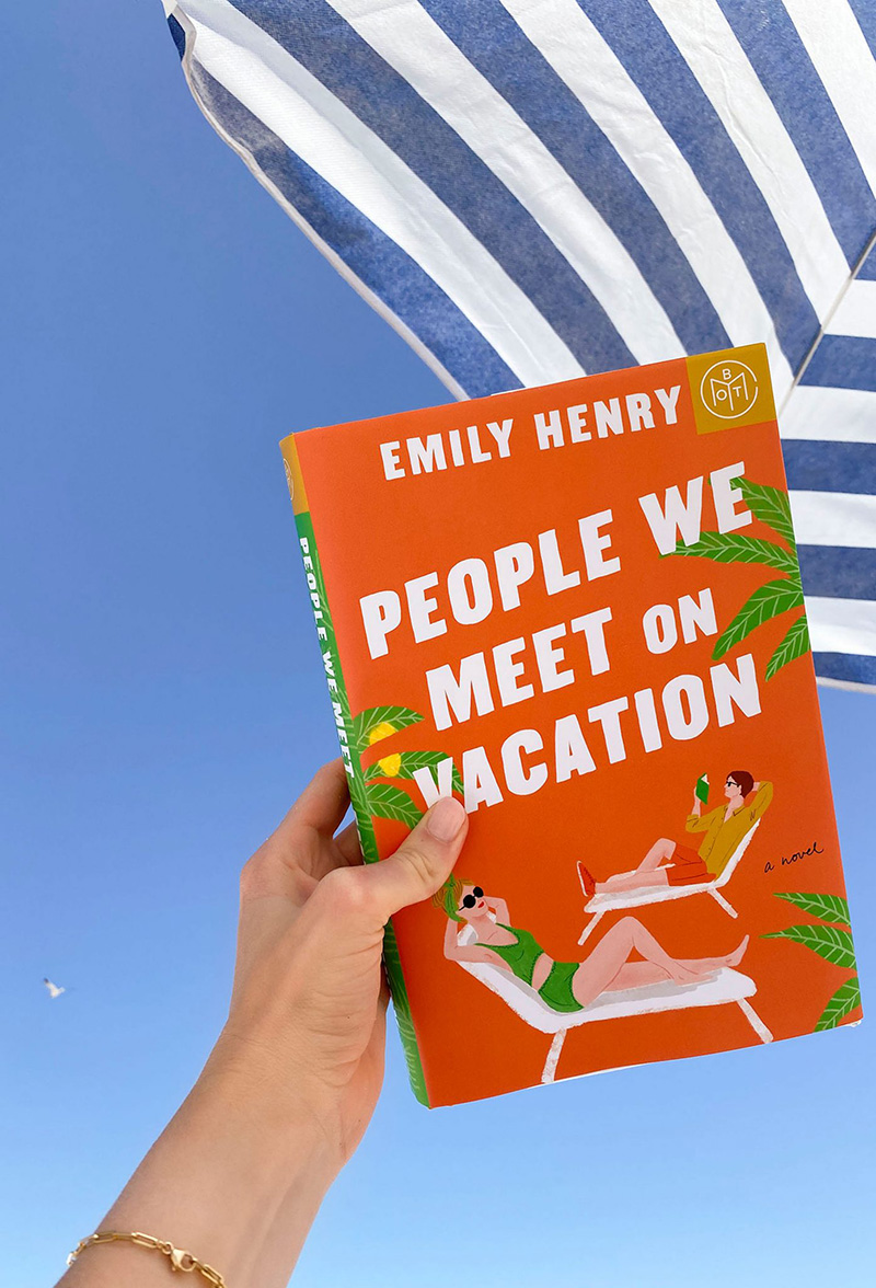 Book cover that reads "People we meet on vacation" being held infront of blue skies and a striped beach umbrella.