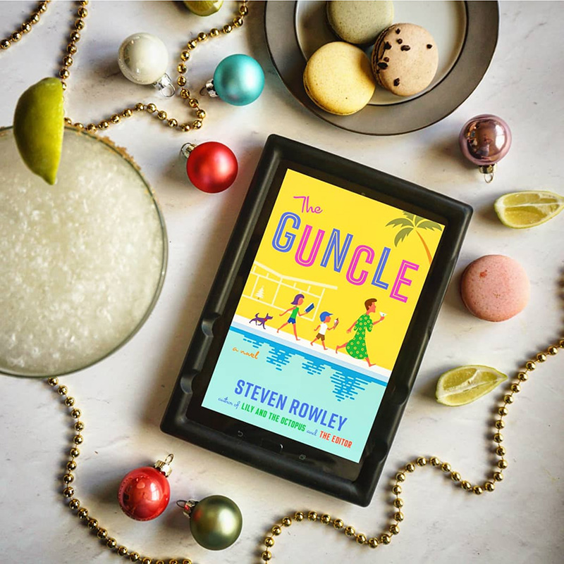 Flatlay with golden beaded garland, Christmas ornament, a margarita, lime slices, and a e-book reader that reads "The Guncle"
